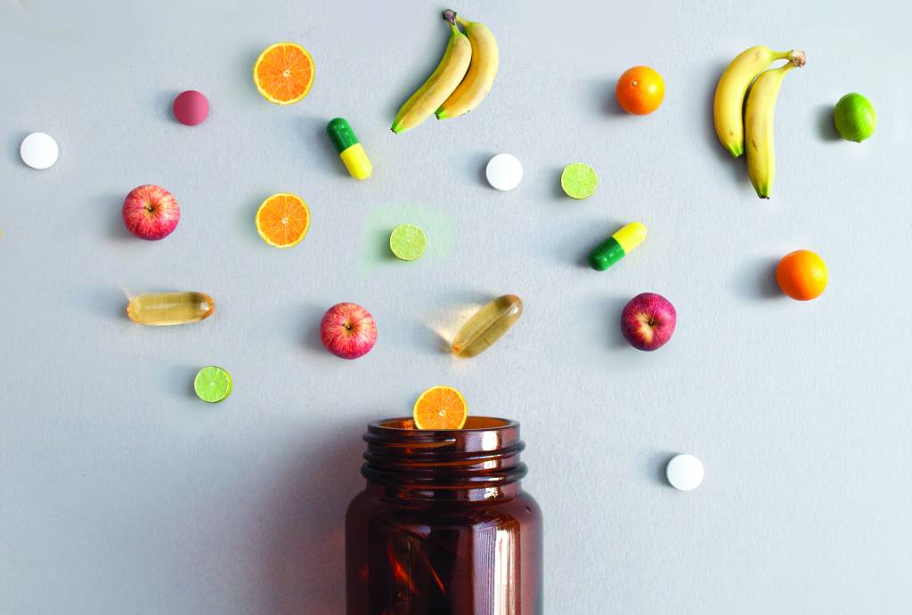 Vitamin pills and capsules alongside apples, bananas, lime and oranges coming out of a medicine jar, nutrition concept