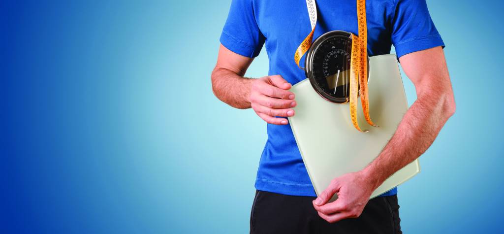 Athletic man with weighing scale and measuring tape hanging dressed in sportswear and isolated blue gradient background.